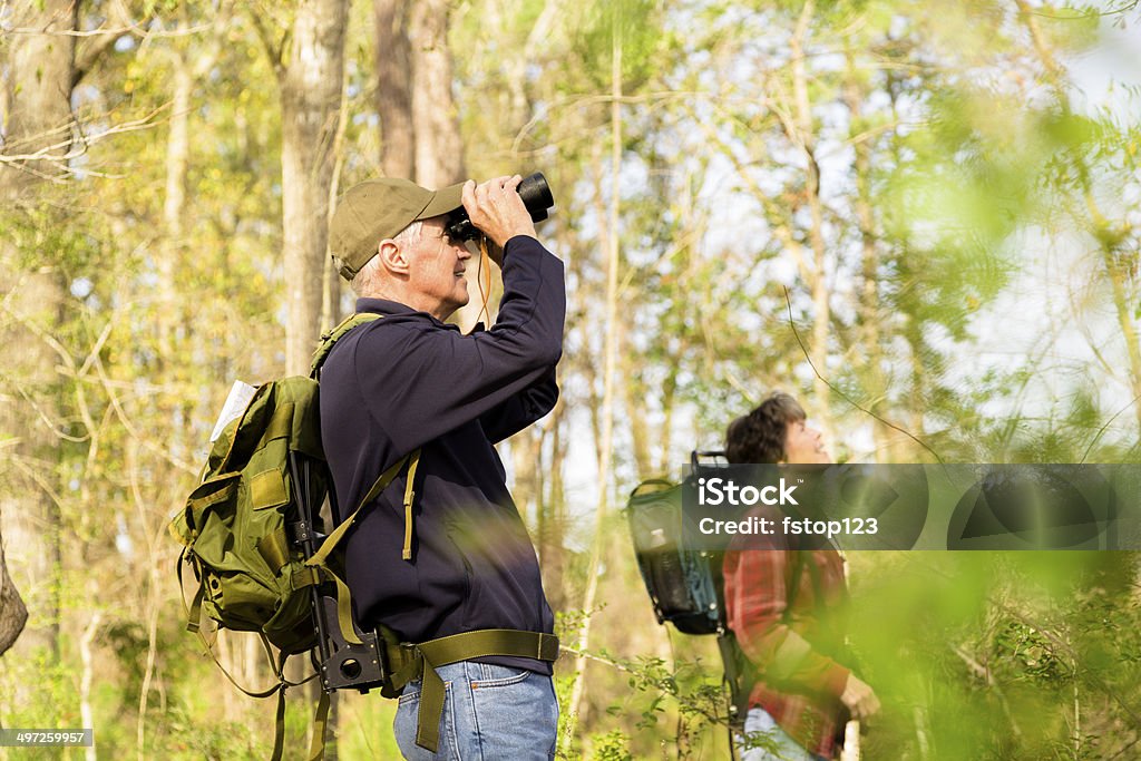 Seniors: Active senior couple outdoors hiking in forest. Nature. Active senior couple outside hiking in a wooded park area of their community. Man uses binoculars to view wildlife.  Bird Watching Stock Photo