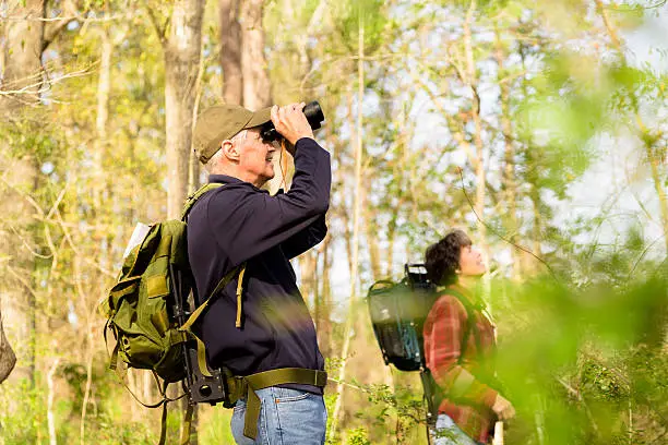 Active senior couple outside hiking in a wooded park area of their community. Man uses binoculars to view wildlife. 