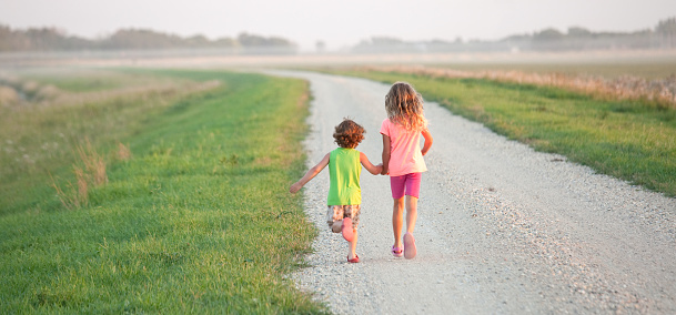 Two young children running down a rural country gravel road. Prairie scene. Elementary aged caucasian children. Panorama