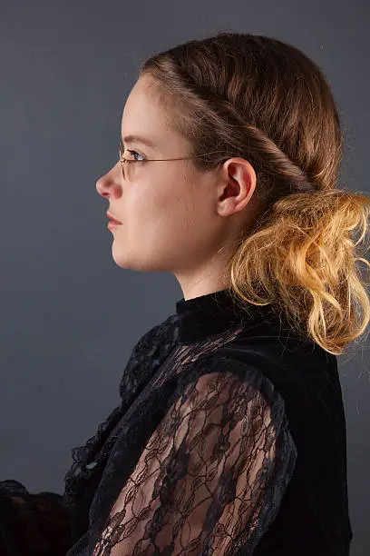 Profile shot of a blonde-haired, bespectacled lady wearing a black, Victorian dress.