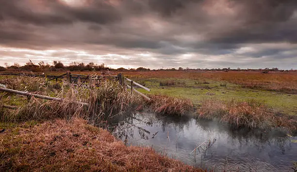 Dramatic autumnal fenland landscape with mist over a drainage ditch spilling onto the land with manacing clouds above