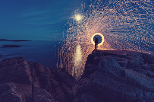 A man sends a spinning shower of sparks into the sea under the light of the Moon.  Long exposure.