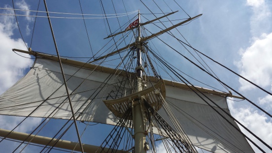 Looking up at an old fashioned, restored sailing ship's main sail hoisted and raised with ropes and rigging around the mast with a blue sky and few clouds in Mystic, Conneticut.