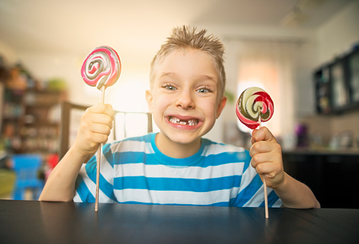 Portrait of a grinning little boy holding two lollipops. The boy's smile is missing couple of teeth, possible because of eating too much candy. 