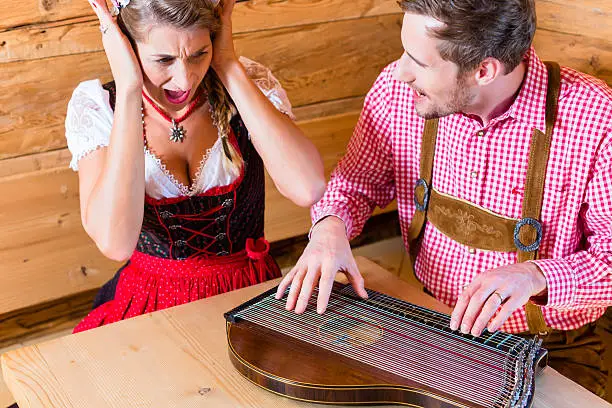 Man playing poorly on zither in mountain hut, his woman in covering her ears with hands