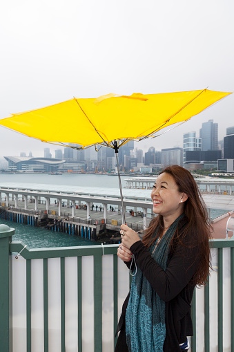 Asian woman with broken umbrella at Hong Kong central piers in the rain. HK convention center in the background.