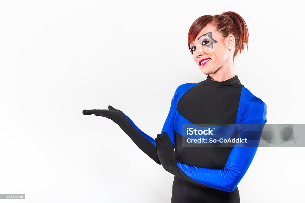 Redhead Woman Presenting In Costume Woman dressed in black and blue body suit stands with her hands out, presenting against a white background. She has red hair and star makeup around her eyes. 20-24 Years Stock Photo