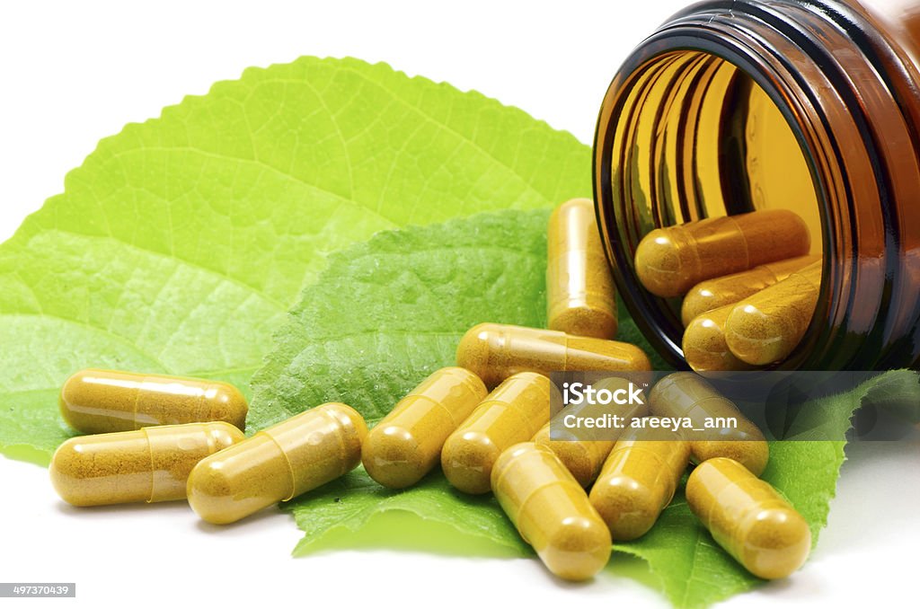 Alternative Medicine Concept. Tumeric Powder in Transparency Hard Gelatin Capsules Contain in Amber Glass Light resistant Bottle Displayed with Green Natural Leaves on White Background. Capsule - Medicine Stock Photo