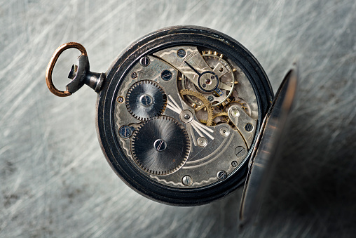 Colour still life of pocket watch with the back cover open showing the intricate and incredibly detailed mechanism. Overhead view with shallow focus, photographed against a distressed metallic background, vertical format with copy space. 