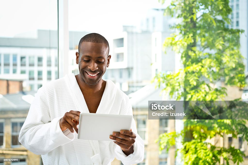 Man in the morning Friendly afro american man wearing white bathrobe standing in her apartment and using a digital tablet. Men Stock Photo