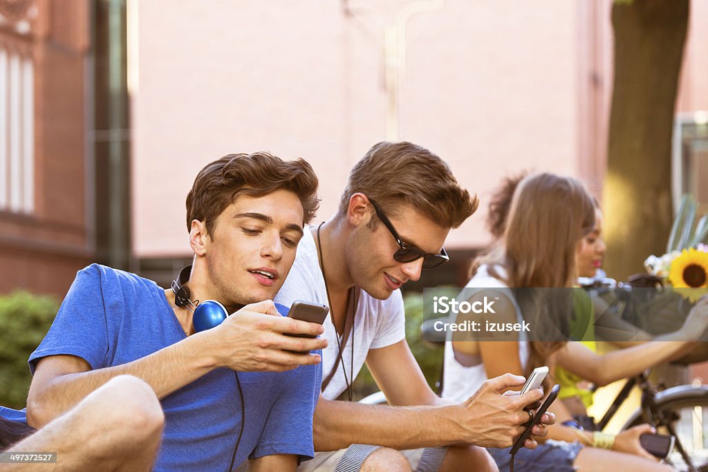 Urban young people Four young urban people sitting outdoors. Focus on two boys using their smart phones. 20-24 Years Stock Photo
