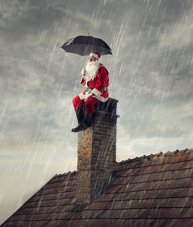 Old man dressed as santa claus sitting on top of a chimney holding his umbrella because it is raining
