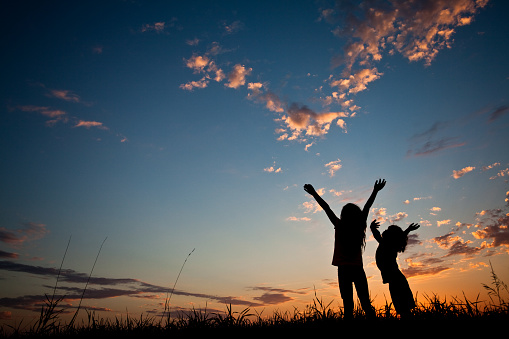 Silhouette of two happy children with their hands raised in worship. Outside. Sunset. Additional themes include praise and worship, kids, celebration, joy, peace, Christianity, religious, balance, wonder, awe, singing, happiness, togetherness, friends, friendship, love, faith, salvation, gratitude, attitude, hope, nature, and outdoors. Children are elementary aged and are unrecognizable. Caucasian models. 