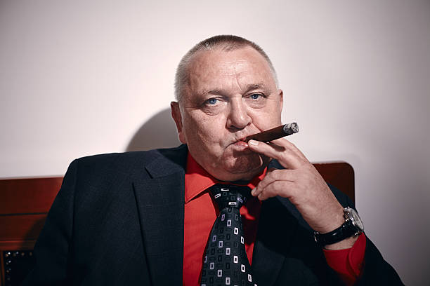 Man with cigar Portrait of serious middle aged businessman wearing black suit, red shirt and wristwatch sitting on old fashioned sofa in office and smoking cigar godfather godparent stock pictures, royalty-free photos & images