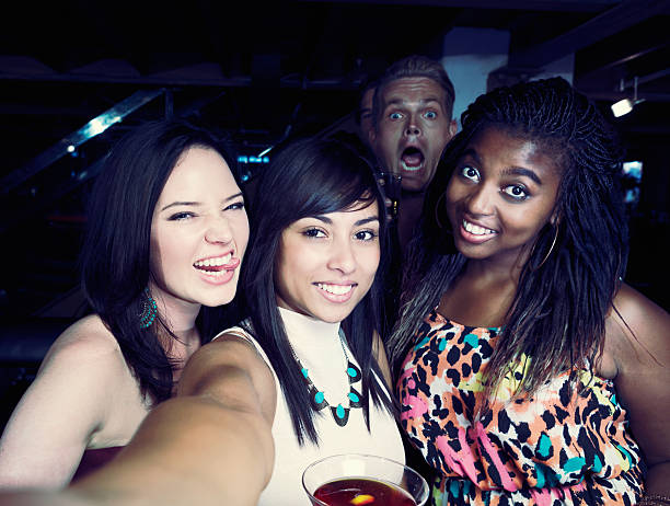 Man plays the fool "photobombing" behind pretty girls taking selfie Three good-looking young women enjoy a night out in a club or bar,  taking selfies ,while a man making silly faces behind them "photo bombs" them. photo bomb stock pictures, royalty-free photos & images