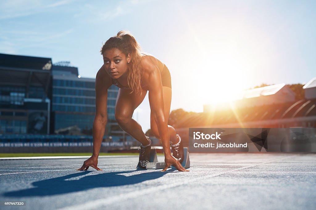 Woman about to start a sprint Confident female athlete in starting position ready for running. Young woman about to start a sprint looking away with bright sunlight from behind. Running Stock Photo