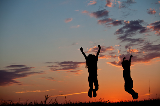 A silhouette of two young children jumping. Horizontal colour image.