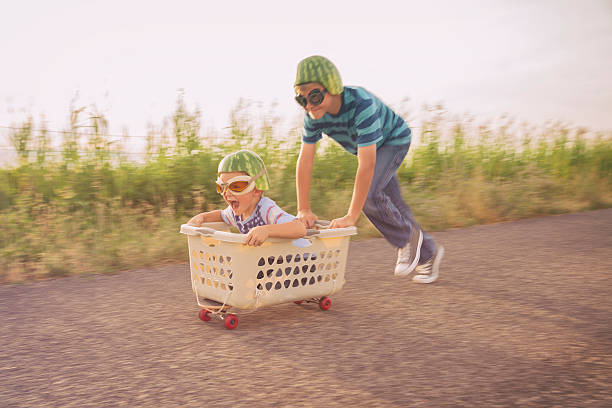 Young Boys Racing on Skateboard Wearing Watermelon Helmets A young boy races his brother in a makeshift go-cart while wearing watermelon helmets and goggles. The two boys smile as they love to be living a life full of adventure. soapbox cart stock pictures, royalty-free photos & images