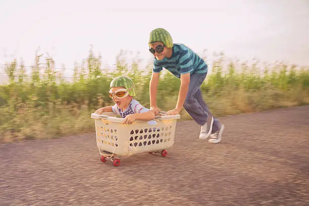 A young boy races his brother in a makeshift go-cart while wearing watermelon helmets and goggles. The two boys smile as they love to be living a life full of adventure.