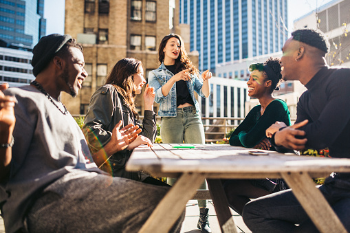 A group of multi-ethnic friends smile and laugh while sitting at a wooden picnic table on a New York City rooftop.  A depiction of friendship and modern urban life in the city.  Horizontal image.