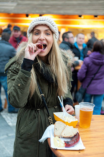 Eating german sausage Front view of beautiful blond cheerful young woman eating in stand with real happiness delicious german sausage in Christmas market in Prague, Czech republic. On the table is fresh good beer. Sausage, mustard and bread are served on paper plate. Cozy and traditional christmas market with christmas decoration are really perfect for spend happy vacations. prague christmas market stock pictures, royalty-free photos & images