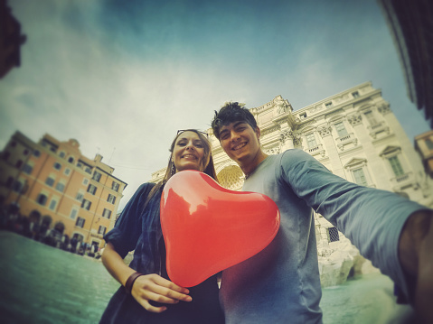Couple taking a selfie with a selfie with heart shaped balloon in front of the restored trevi fountain in Rome