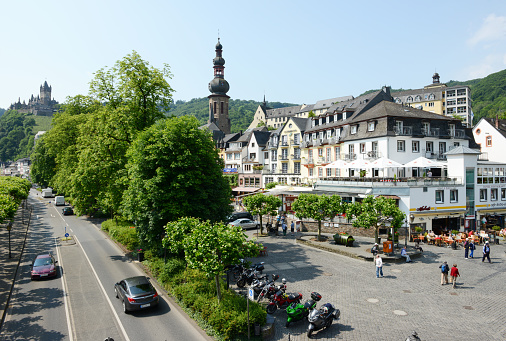 Cochem, Germany - June 2, 2014: People visit the town Cochem (Rhineland Palatinate, Germany) in Mosel Valley. In front cars driving on road leading along the mosel river. People visiting  the old town part small resuatrants and bars. Cars and motorcycles parked on the square.