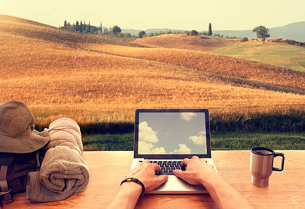 Digital nomad using laptop computer from an hillside italian town Digital nomads are people who can work from anywhere using a laptop and an internet connection: from an hillside italian town in Tuscany a young male traveller is typing on his laptop computer on a wooden table, with a travel backpack and a mug with coffee. On the background a beautiful panorama of hills covered by corn fields and trees at sunset. Laptop display reflects sky with clouds. digital nomad stock pictures, royalty-free photos & images