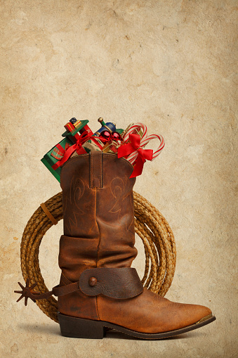 A cowboy boot with a spur attached is stuffed with Christmas presents, a toy train, and candy canes on a warm textured background. There is a lasso behind cowboy boot. Clipping path included.