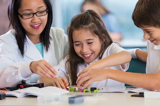 Young adult Asian female science teacher in private elementary school is assisting male and female Hispanic elementary age students. Children are building a robotic model using wires and parts. Students are smiling while learning about technology. They are wearing private school uniforms.