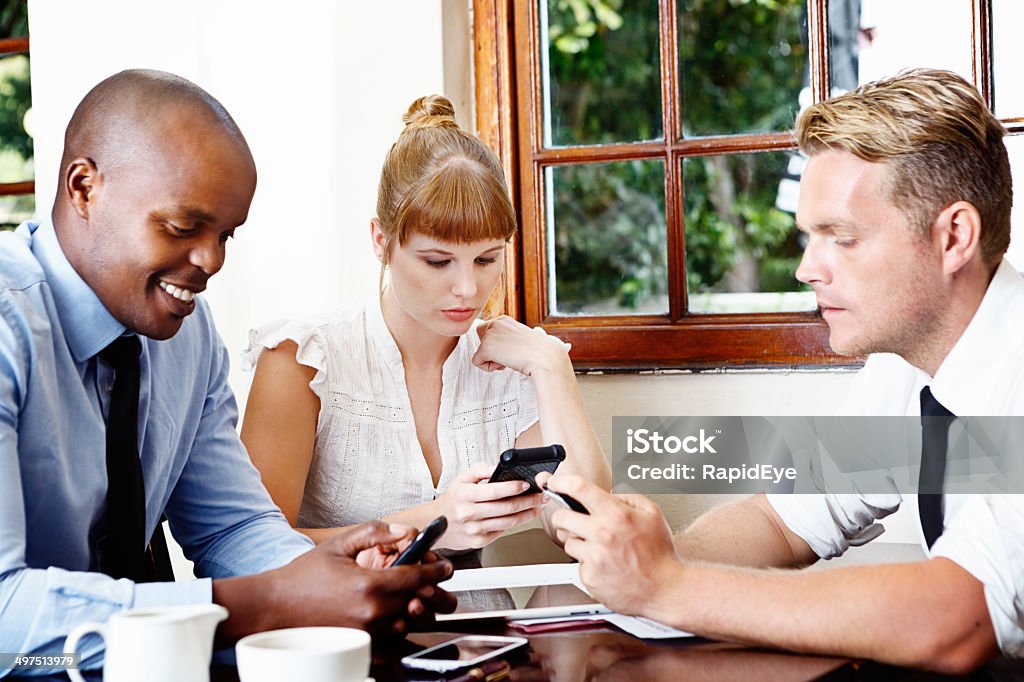 Three young business people in meeting, intent on their cellphones Three young business people, two men and a woman, sit over coffee at a cafe, looking down at their cellphones rather than talking to each other directly. It's the modern way to communicate! Adult Stock Photo