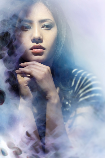 Indoor image of a beautiful serene Asian late teen or young woman sitting behind a murky glass window and looking away at something through it with blank expression. Vertical image with copy space and selective focus.