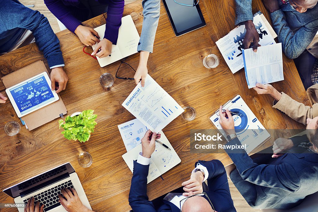 Papers are passed in a business meeting A group of multi-ethnic people gathers around a brown wooden table and works on a project.  They have papers, laptops and tablets. Meeting Stock Photo