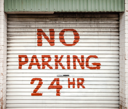 Hand-painted red sign on grimy cream colored lane garage roller door stating NO PARKING 24 HR