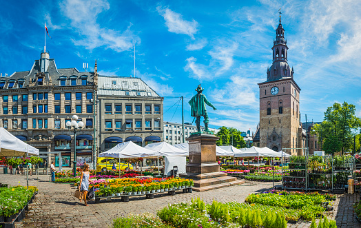 Oslo, Norway - July 12, 2014: Woman walking between market stalls selling green plants and colourful flowers in Stortorvet, the Grand Plaza in the hear of downtown Oslo, Norway. Composite panoramic image created from six contemporaneous sequential photographs. 