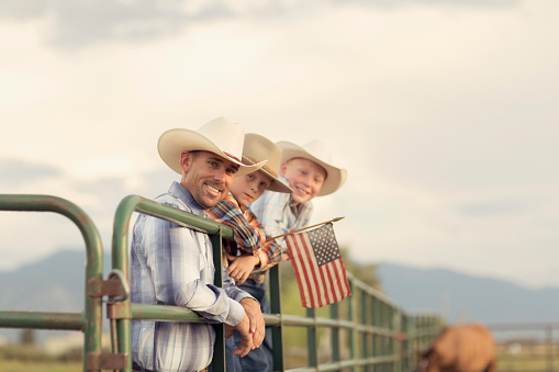 An American father and sons in western wear take a portrait on their farm in Utah. They are quintessential cowboys and cowgirls standing on a ranch gate holding a USA flag.