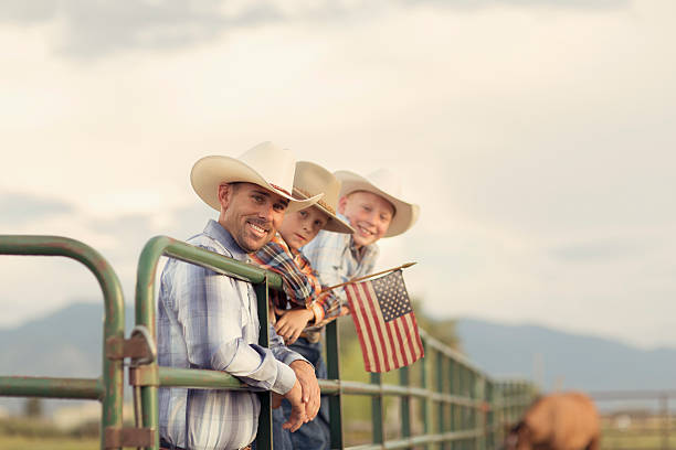 American West Boys dressed Holding USA Flag An American father and sons in western wear take a portrait on their farm in Utah. They are quintessential cowboys and cowgirls standing on a ranch gate holding a USA flag. rural main street stock pictures, royalty-free photos & images