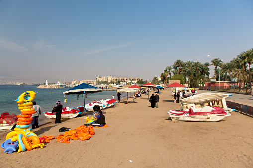 Aqaba , Joardan - January 27, 2015:  Public beach in Aqaba. Some tourist boats and motorboats in the middle distance. Palm trees and hotels in the background.