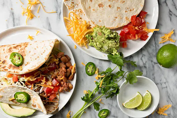 An overhead close up horizontal photograph of a couple of soft shell tacos on a plate, extra flour tortillas, guacamole, tomatoes and shredded cheddar cheese.