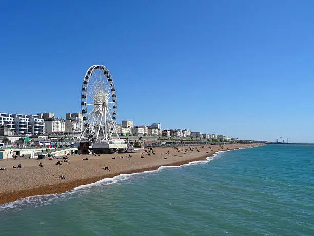 Photo showing the big wheel attraction at Brighton beach, next to the pier.  The beach is pictured in the morning, when the sun is shining and tourists are sunbathing on the sandy shingle / pebbles.