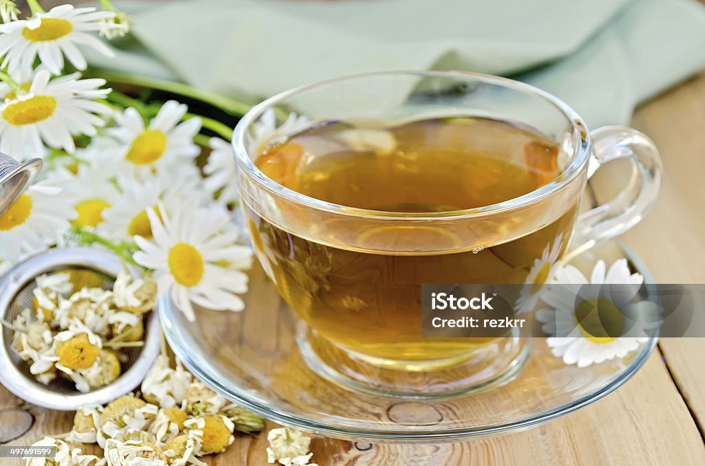 Herbal chamomile tea in cup with strainer on board Herbal tea in a glass cup, metal sieve with dry chamomile flowers, fresh flowers, daisies, green cloth on a background of wooden boards Alternative Therapy Stock Photo