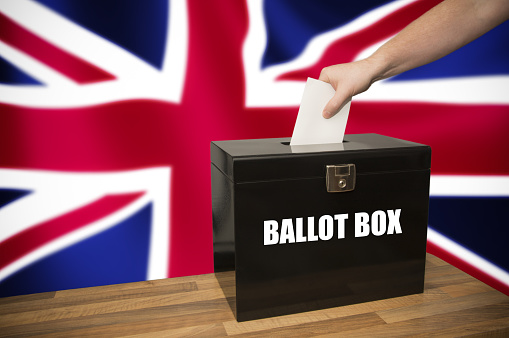 A hand casting a vote in a ballot box against the flag of the United Kingdom. General elections, local elections, EU referendum and other votes.