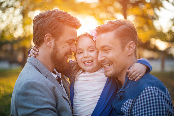 Gay Parents with daughter Young gay parents with their daughter having fun in park. Parents holding girl in arms. Enjoying in beautiful sunset. Caucasian ethnicity. homosexual stock pictures, royalty-free photos & images