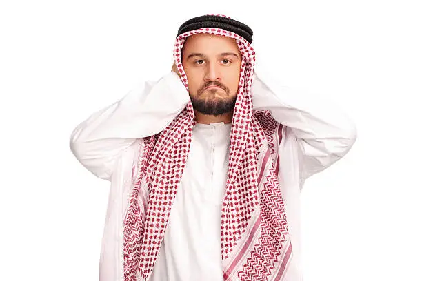 Reluctant male Arab covering his ears with his hands and looking at the camera isolated on white background