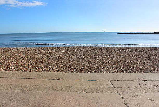 Photo showing a concrete seaside promenade in the foreground, while behind is a shingle beach, sand, sea and a blue summer's sky sky.