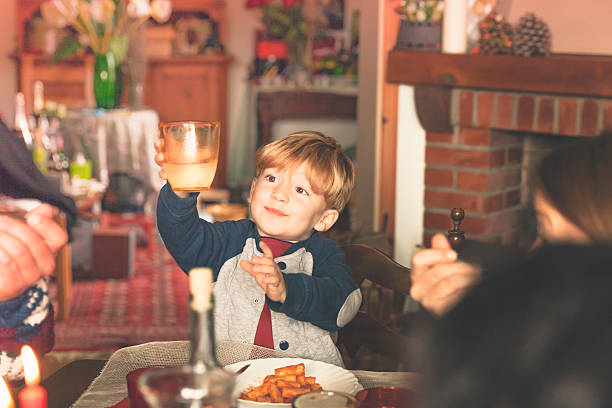A young blonde boy is holding his glass to cheer an happy new year at table while eating pasta