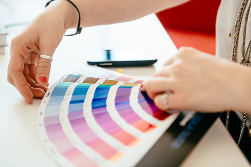 Close-up shot of designers hands holding a color fan and choosing the right one. she is sitting in her office on a table.