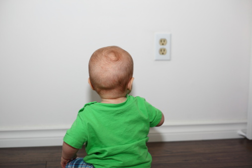 A close-up image of a cute Caucasian baby boy sitting on the floor and looking at the electrical outlet on the wall. Safety hazards for babies at home. Baby hazards. See more in my profile.