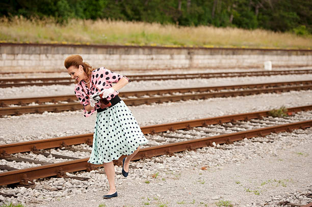 Beautiful and smiling adult woman with long brown hair in retro style 1950, wearing doted blue skirt, pink blouse and white lace gloves. She holding an old camera in leather pouch. She walking and dancing on a train station.