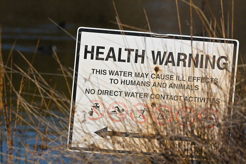 Warning sign photographed on a small lake surrounded by walking paths in an urban area. HEALTH WARNING - THIS WATER MAY CAUSE ILL EFFECTS TO HUMANS AND ANIMALS - NO DIRECT WATER CONTACT ACTIVITIES